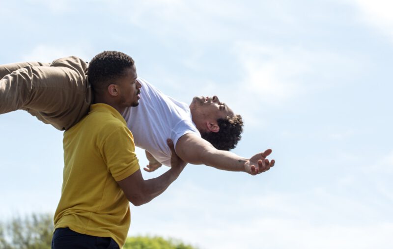 Two circus performers. One holding the other on his back over his shoulder.